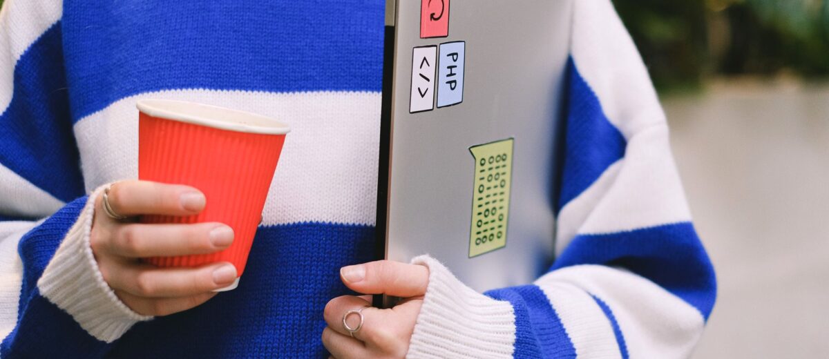 Person wearing striped sweater holding laptop with stickers and a red coffee cup indoors.