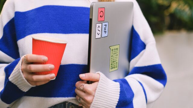 Person wearing striped sweater holding laptop with stickers and a red coffee cup indoors.