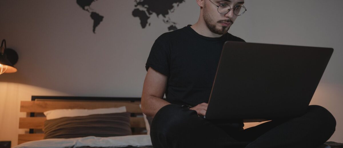 A man sits on a bed with a laptop, working remotely, with a world map and pizza plate nearby.