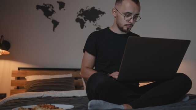 A man sits on a bed with a laptop, working remotely, with a world map and pizza plate nearby.
