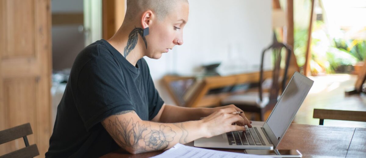 Bald woman with tattoos working on a laptop in a home office. Natural light creates a serene workspace.