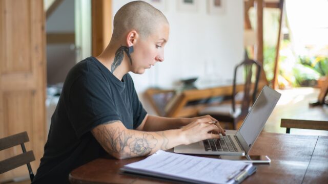 Bald woman with tattoos working on a laptop in a home office. Natural light creates a serene workspace.