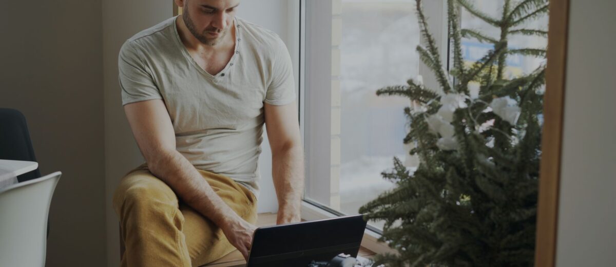 Casual man using laptop indoors by a window, with camera and plant nearby.