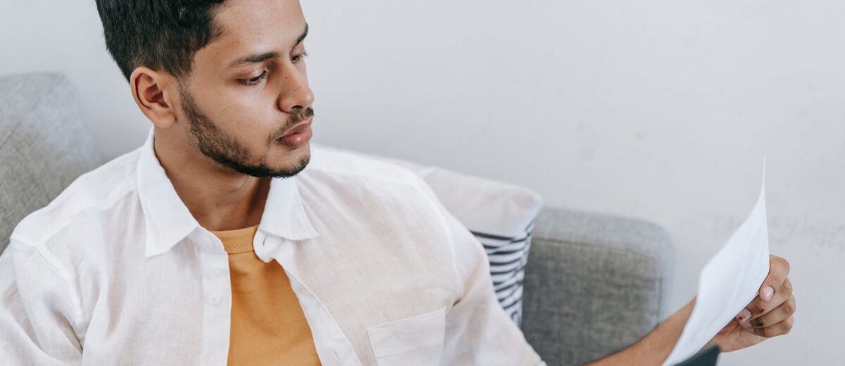 Concentrated young ethnic male wearing casual outfit reading document and browsing netbook while resting on cozy couch