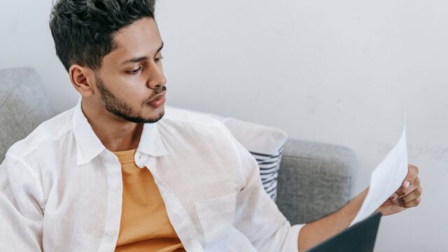 Concentrated young ethnic male wearing casual outfit reading document and browsing netbook while resting on cozy couch
