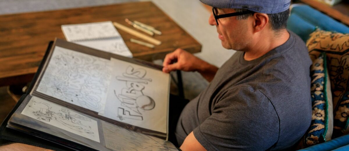 A man sits on a sofa reviewing his art portfolio filled with various illustrations.