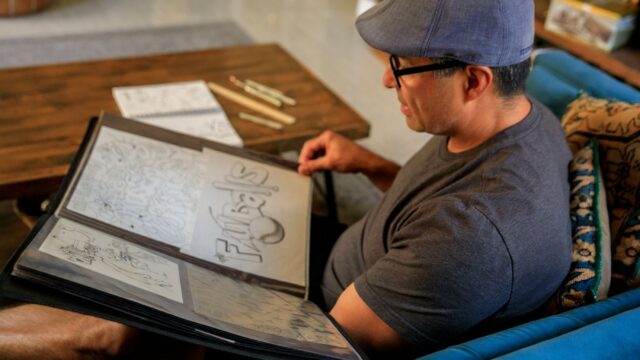 A man sits on a sofa reviewing his art portfolio filled with various illustrations.