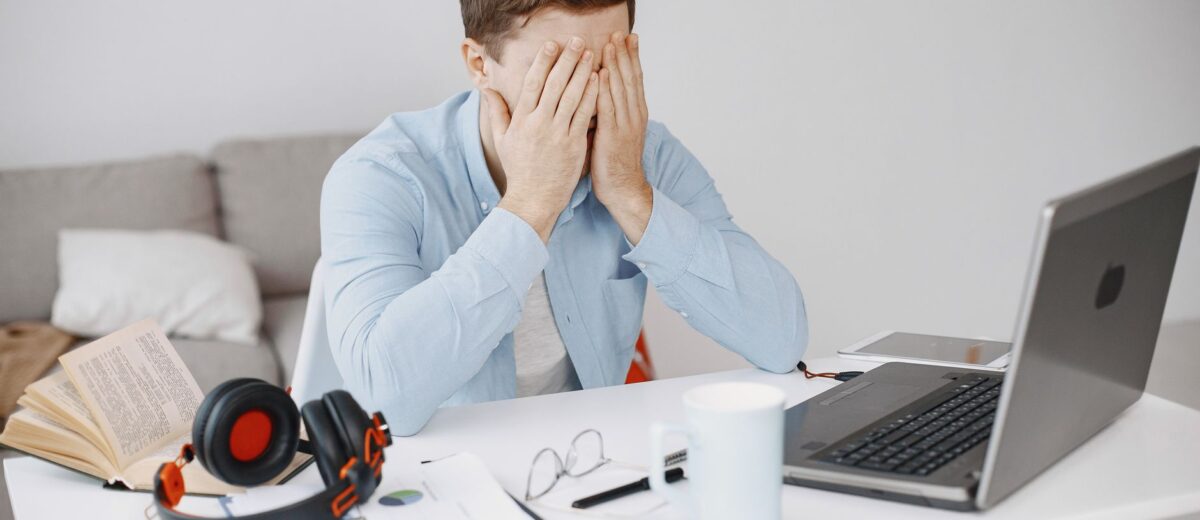 Stressed man at home office desk holding head in frustration over laptop work.