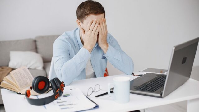 Stressed man at home office desk holding head in frustration over laptop work.