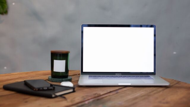 A modern workspace featuring a blank laptop screen, notebook, and candle on a wooden table.