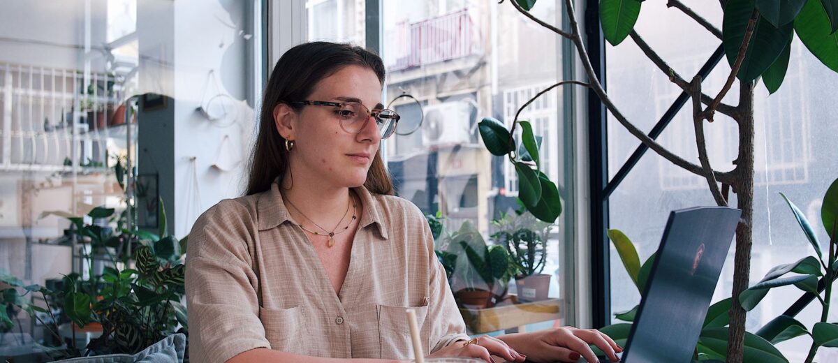 Young woman using laptop in a stylish café with plants and natural light. Ideal for business or lifestyle themes.