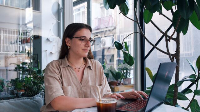 Young woman using laptop in a stylish café with plants and natural light. Ideal for business or lifestyle themes.