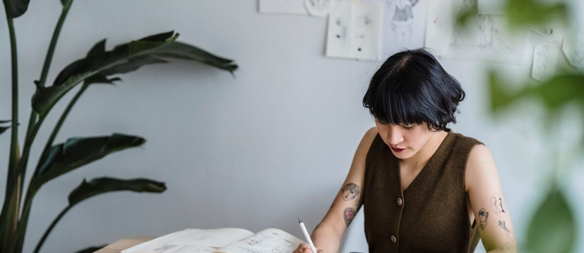 Concentrated Asian female artist sitting at table with graphics tablet and drawing sketches in creative workspace