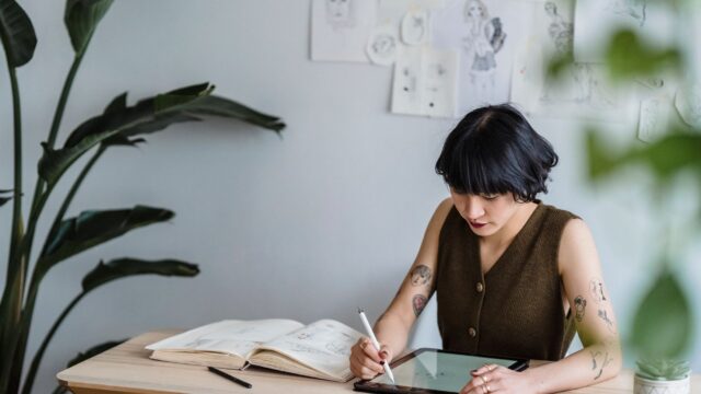 Concentrated Asian female artist sitting at table with graphics tablet and drawing sketches in creative workspace