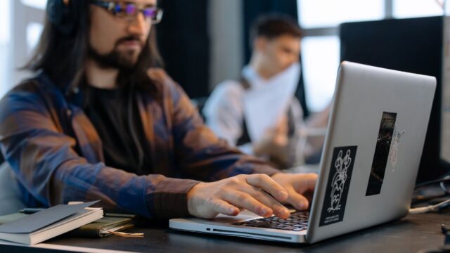 A man wearing headphones focused on typing at his laptop in a contemporary office setting.